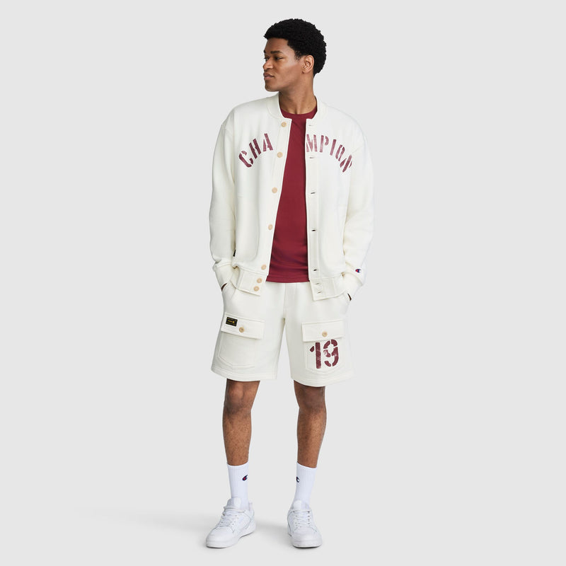 A person wearing a cream-colored Champion Fleece Baseball Jacket with maroon lettering over a maroon t-shirt and matching cream cargo shorts with a maroon number 19 patch.
