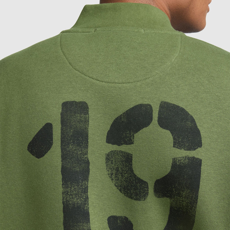 A person wearing a Champion Fleece Baseball Jacket in green, featuring a large, black, distressed-style number 19 printed on the upper back, evoking a vintage athletic vibe reminiscent of a classic varsity jacket. Only the upper back and neck are visible.