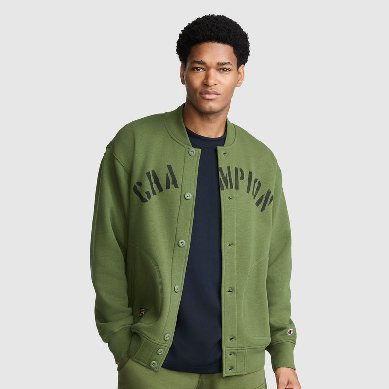 A person wearing a Champion Fleece Baseball Jacket in olive green, featuring black “CHAMPION” lettering, over a black shirt stands against a plain light gray background.