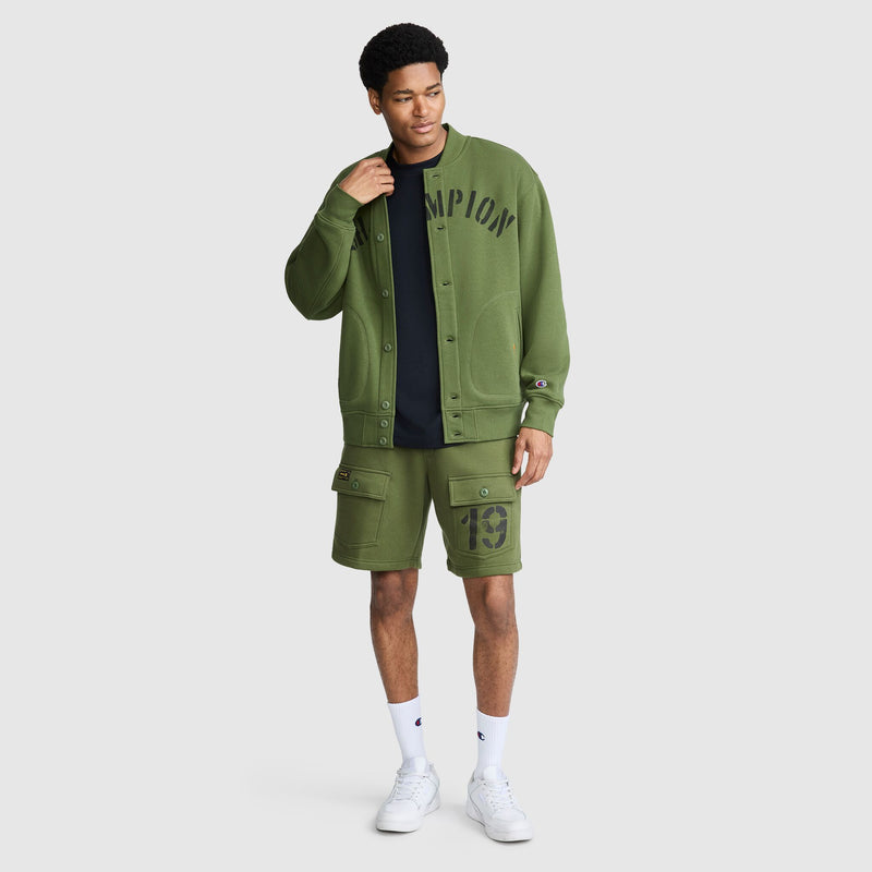 A man stands against a plain light background wearing a green Champion Fleece Baseball Jacket, coordinating green shorts featuring the number 19, a black T-shirt, white socks, and white sneakers. The look channels a vintage athletic vibe with its classic varsity jacket styling.