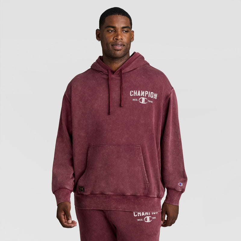 A man wears a burgundy Mineral Wash Pullover Hoodie, C Patch Logo with a logo graphic on the chest and a small emblem on the sleeve.