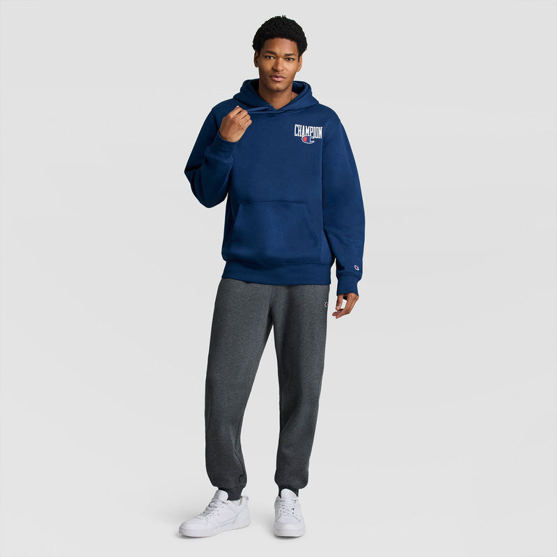 A man wearing a navy blue Fleece Hoodie, Left Chest Graphic with a white graphic on the chest, grey sweatpants, and white sneakers.