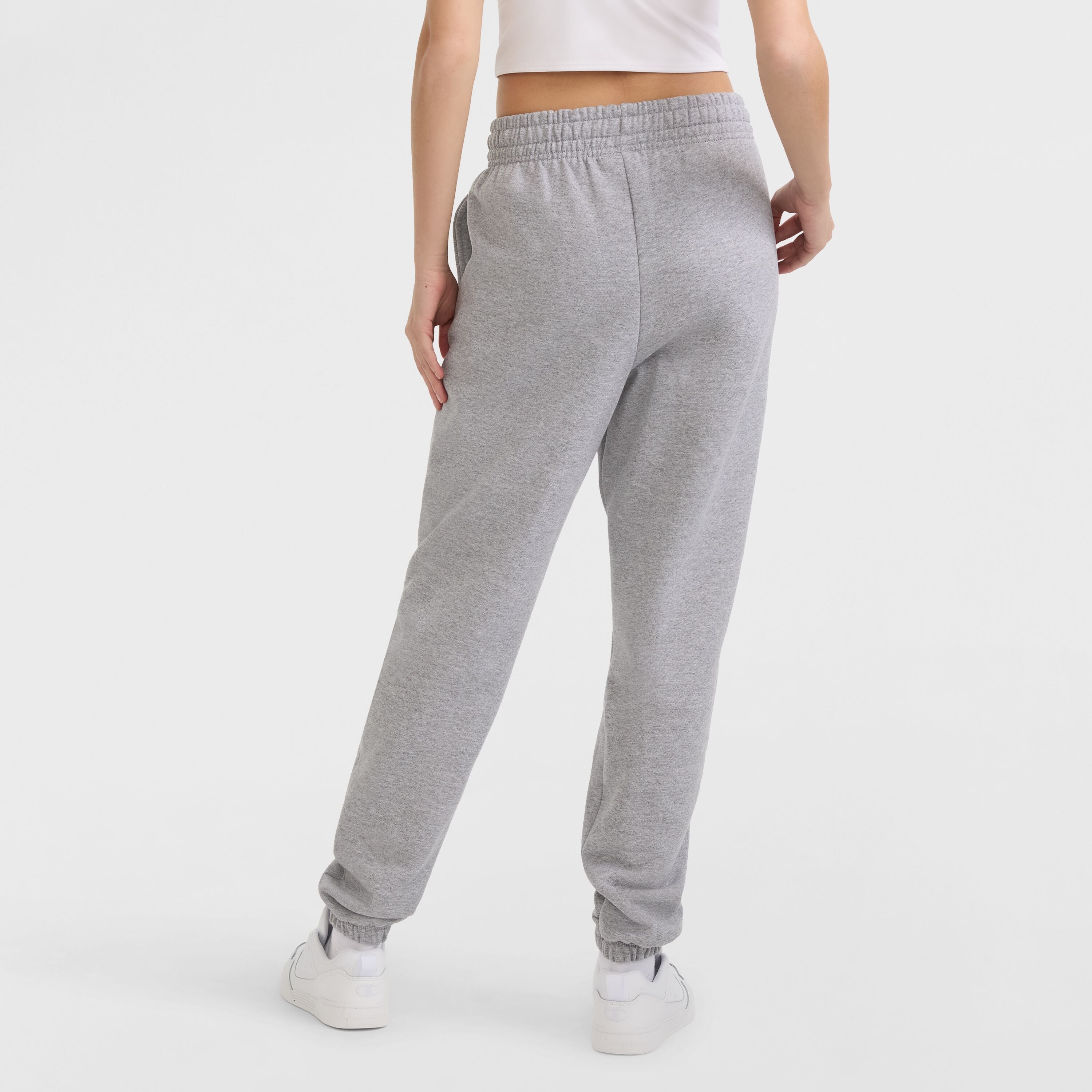 A person wearing Champion Powerblend Oversized Sweatpants with a C Logo, a white cropped top, and white sneakers stands with their back to the camera against a plain white background.