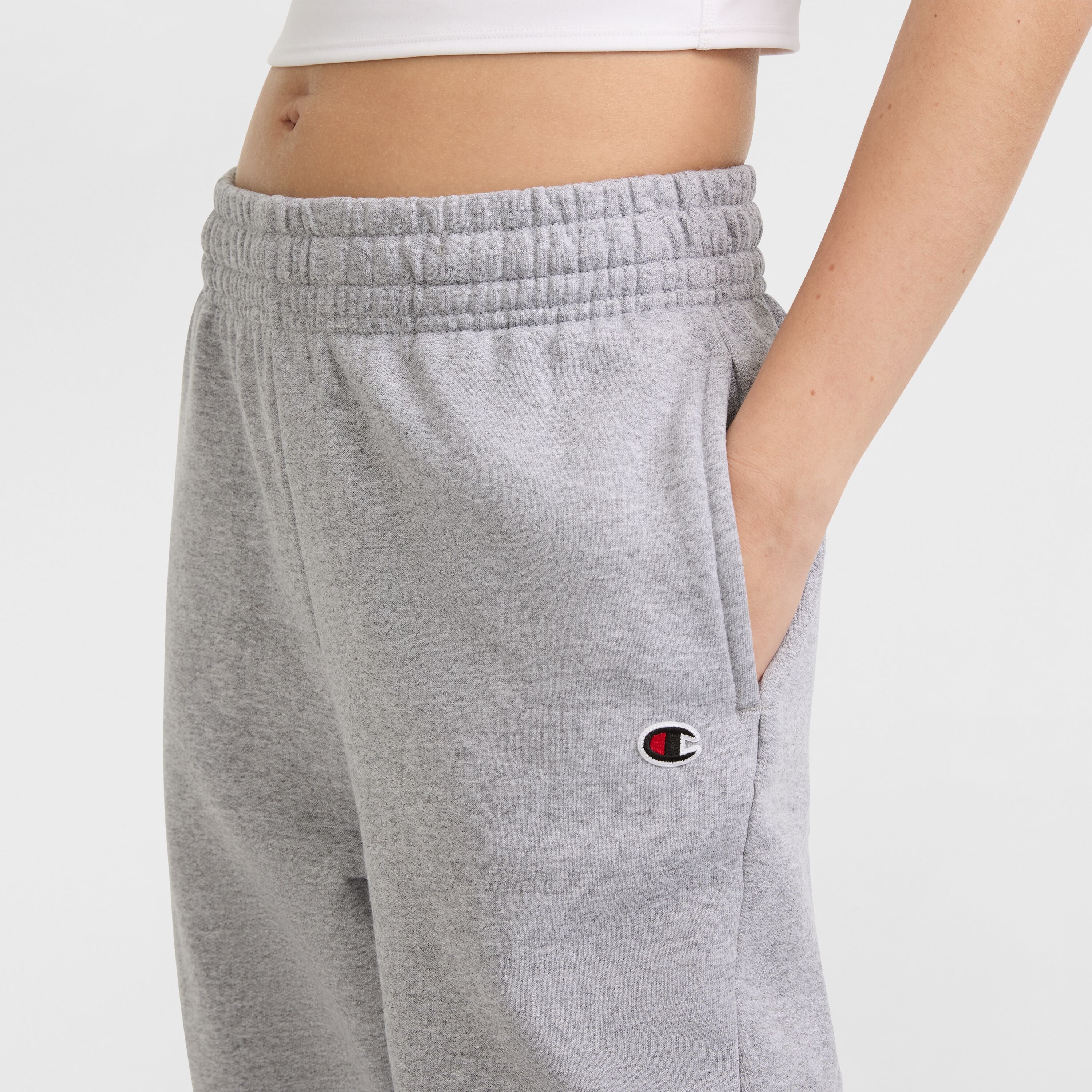 A person wears Champion Powerblend Oversized Sweatpants featuring a C logo on the left hip, elastic waistband, light gray color, one hand in the pocket, and part of a white crop top showing.