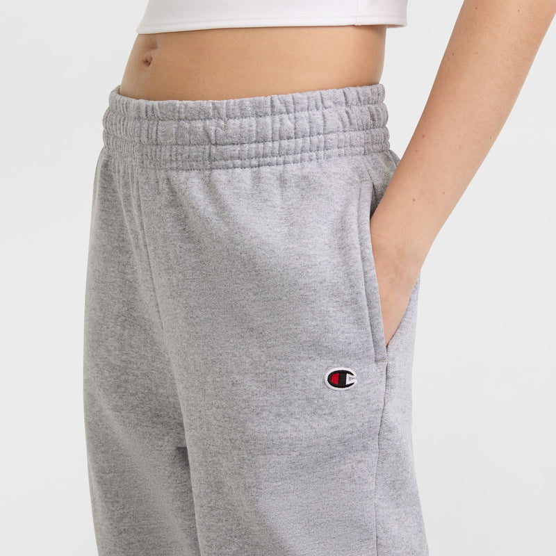A person wears Champion Powerblend Oversized Sweatpants featuring a C logo on the left hip, elastic waistband, light gray color, one hand in the pocket, and part of a white crop top showing.