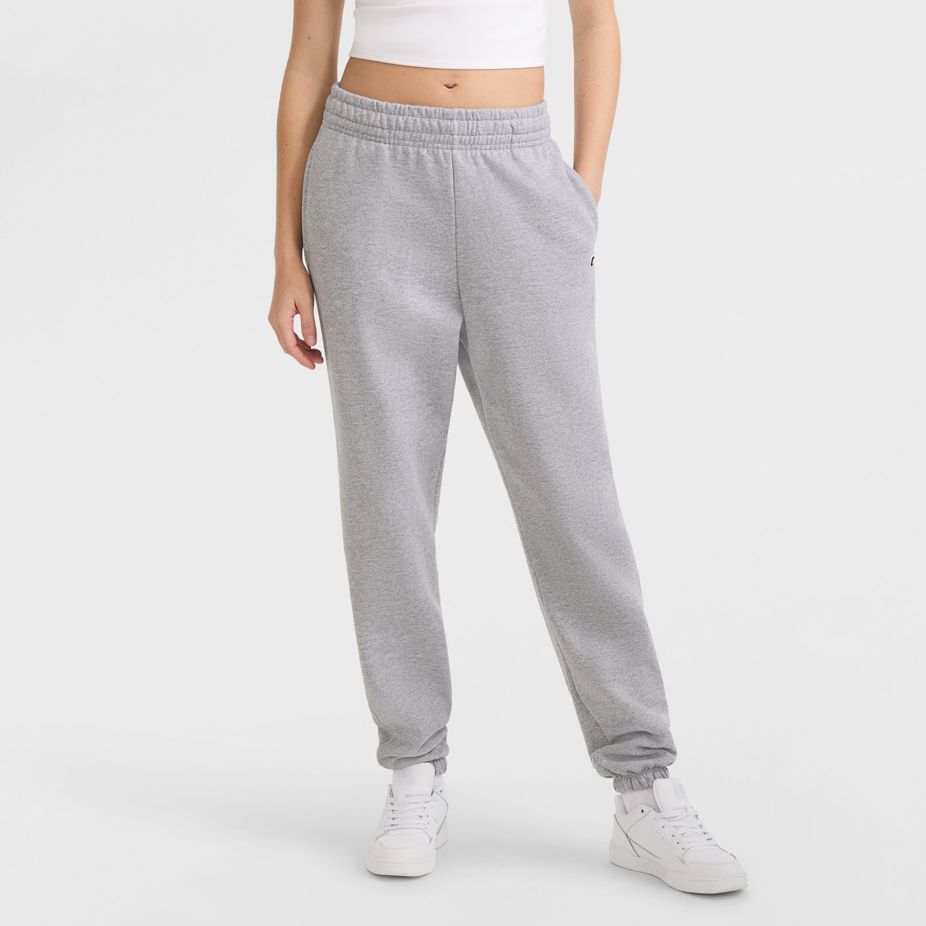 A person stands against a plain white background wearing a white crop top, Champion Powerblend Oversized Sweatpants with C Logo in light gray, and white sneakers. Their head is not visible.