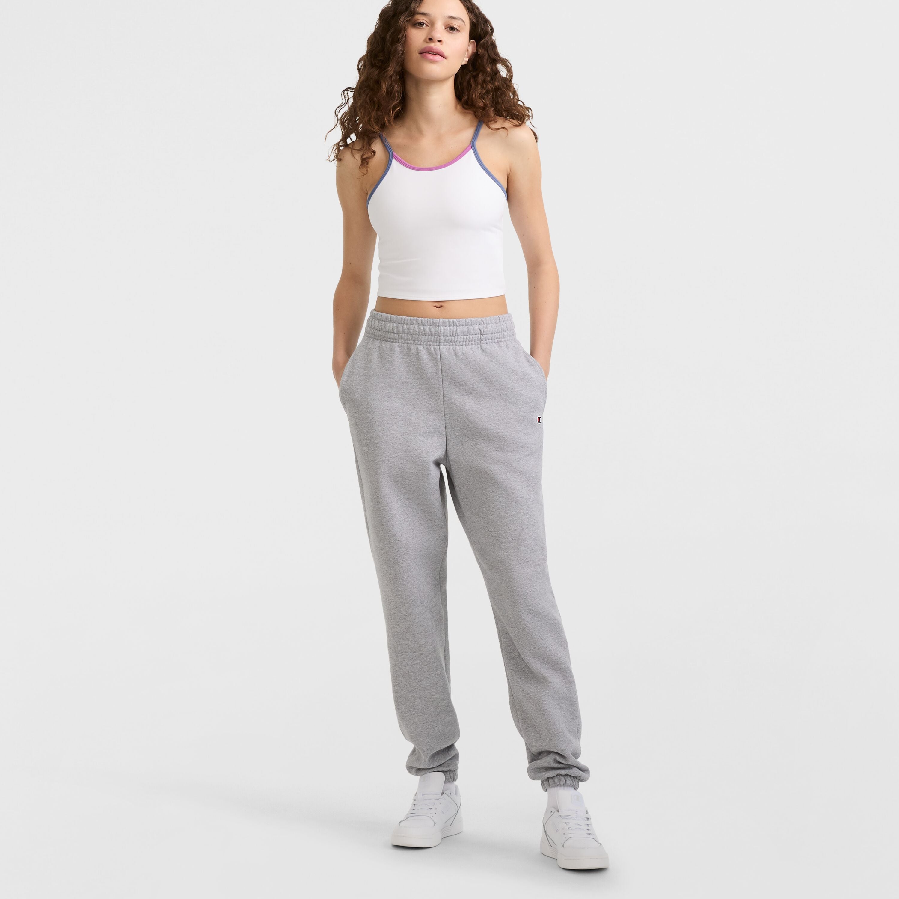 A person with curly hair, wearing a white crop tank top with purple trim, Champion Powerblend Oversized Sweatpants (C Logo), and white sneakers stands with hands in pockets against a light background.