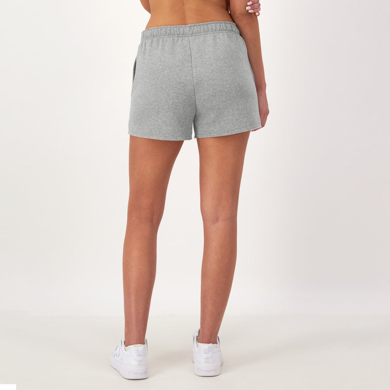 A person stands on a white background, shown from the back, wearing Champion 3" Powerblend Shorts with the C Logo in light gray, paired with white sneakers and a crop top. Their arms rest relaxed by their sides.