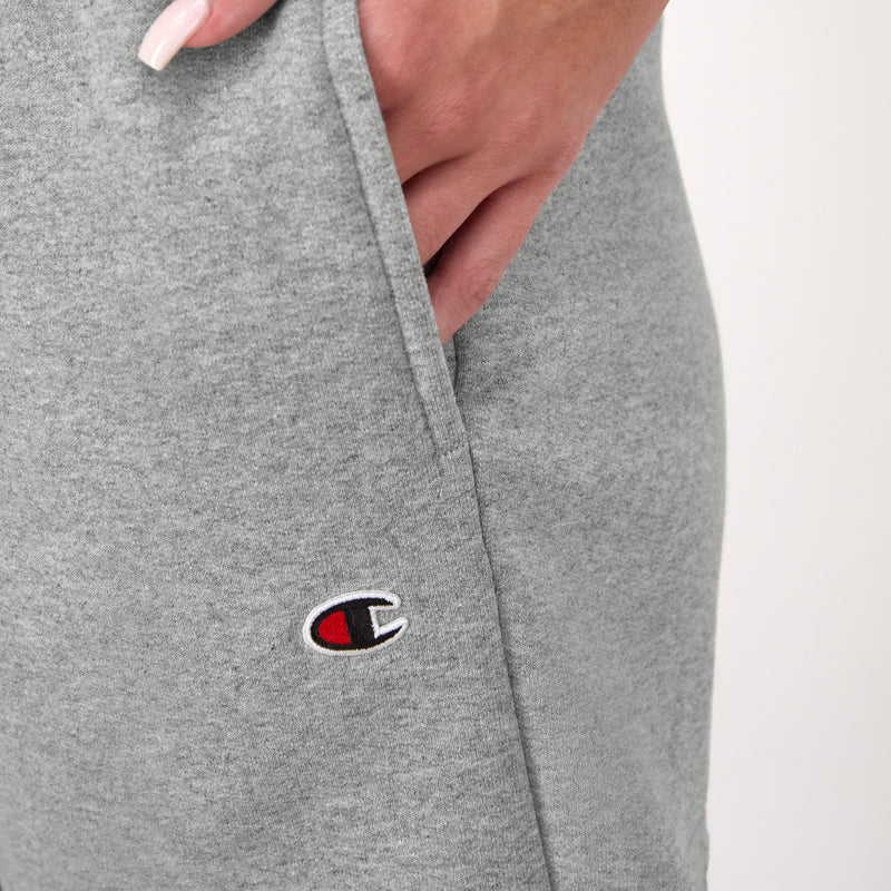 A close-up of a hand with manicured nails resting in the pocket of light grey Champion 3" Powerblend Shorts, featuring the C Logo embroidered near the pocket.