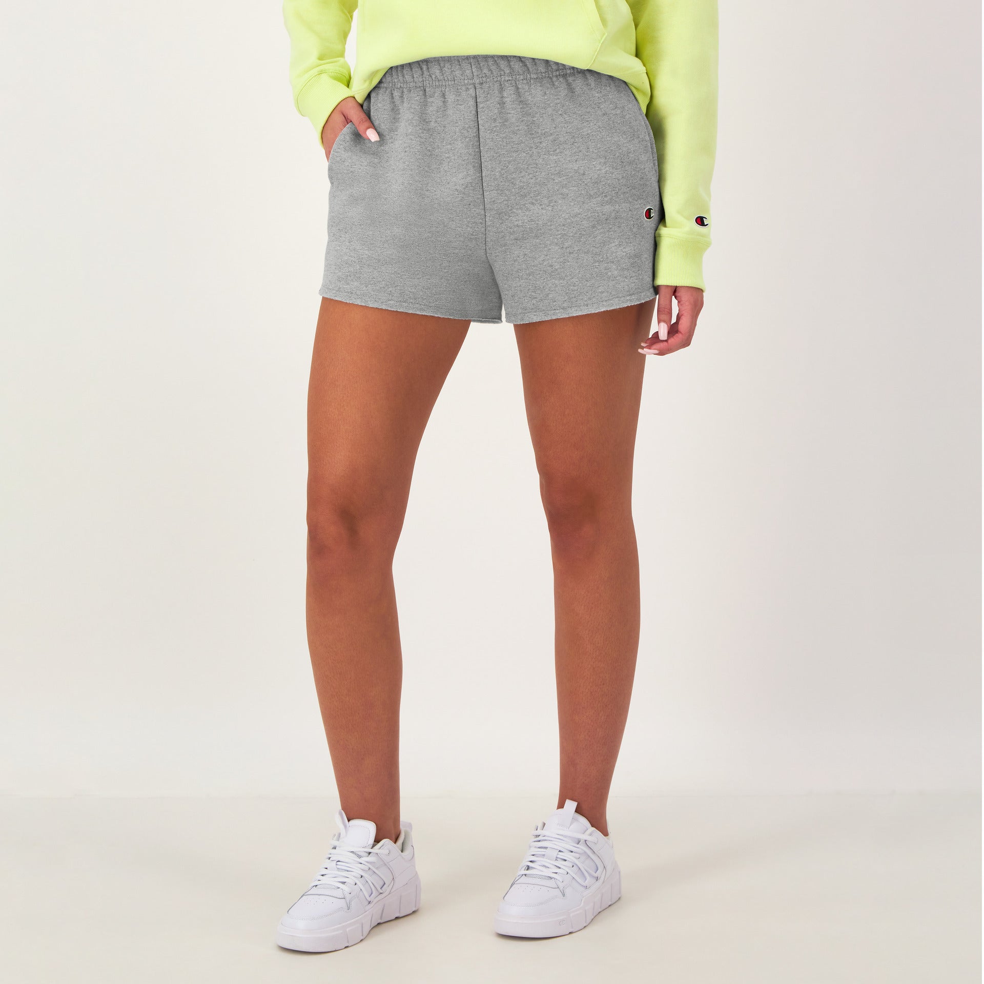 A person in a neon yellow sweatshirt, white sneakers, and Champion 3" Powerblend Shorts, C Logo in gray stands against a plain light background with one hand in their pocket.