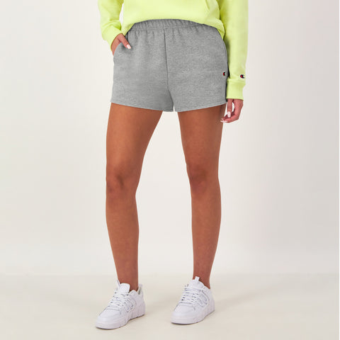 A person in a neon yellow sweatshirt, white sneakers, and Champion 3" Powerblend Shorts, C Logo in gray stands against a plain light background with one hand in their pocket.