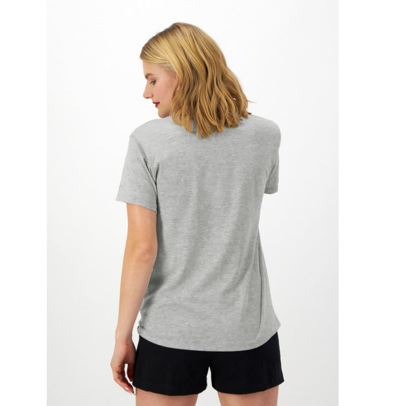 A woman with shoulder-length blonde hair stands with her back to the camera, wearing a light gray Champion Classic T-Shirt, Script Logo, and black shorts against a plain white background.