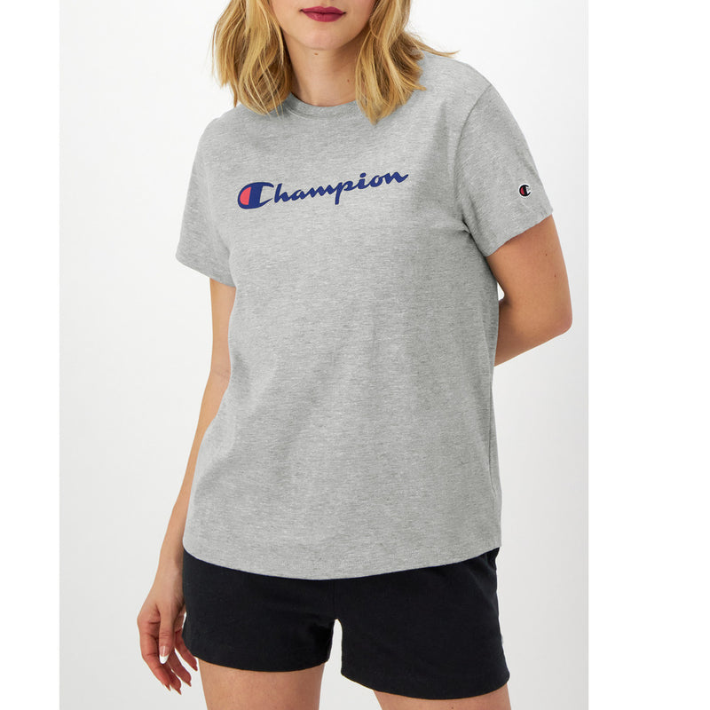 A person wears a light gray Champion Classic T-Shirt with the script logo in blue and red on the chest and black shorts, standing against a plain white background.