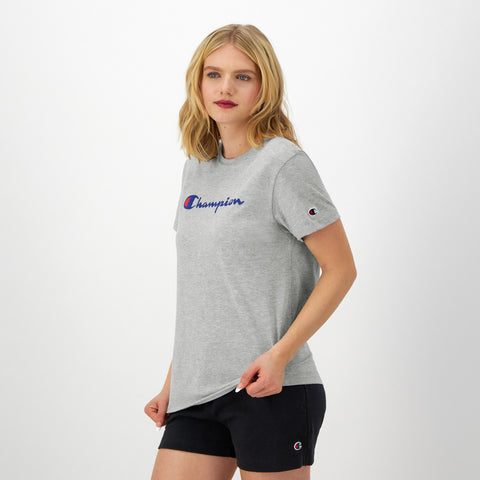 A blonde woman wears the Champion Classic T-Shirt with script logo and black Champion shorts, standing against a white background and gazing to the side with a neutral expression.