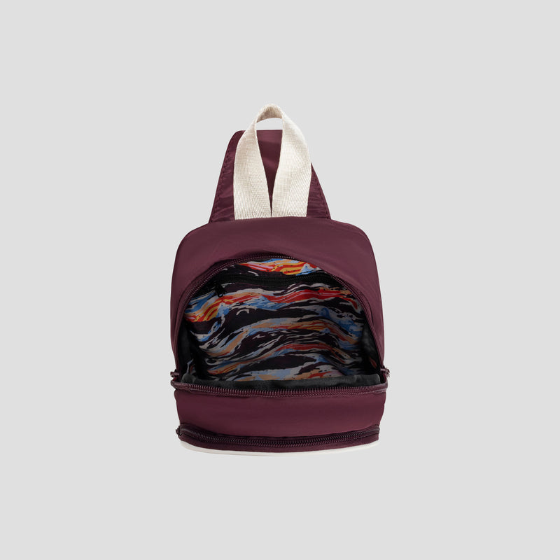 A maroon Champion Oracle Sling Pack with a white handle sits open, revealing a colorful abstract-patterned interior on a plain gray background.