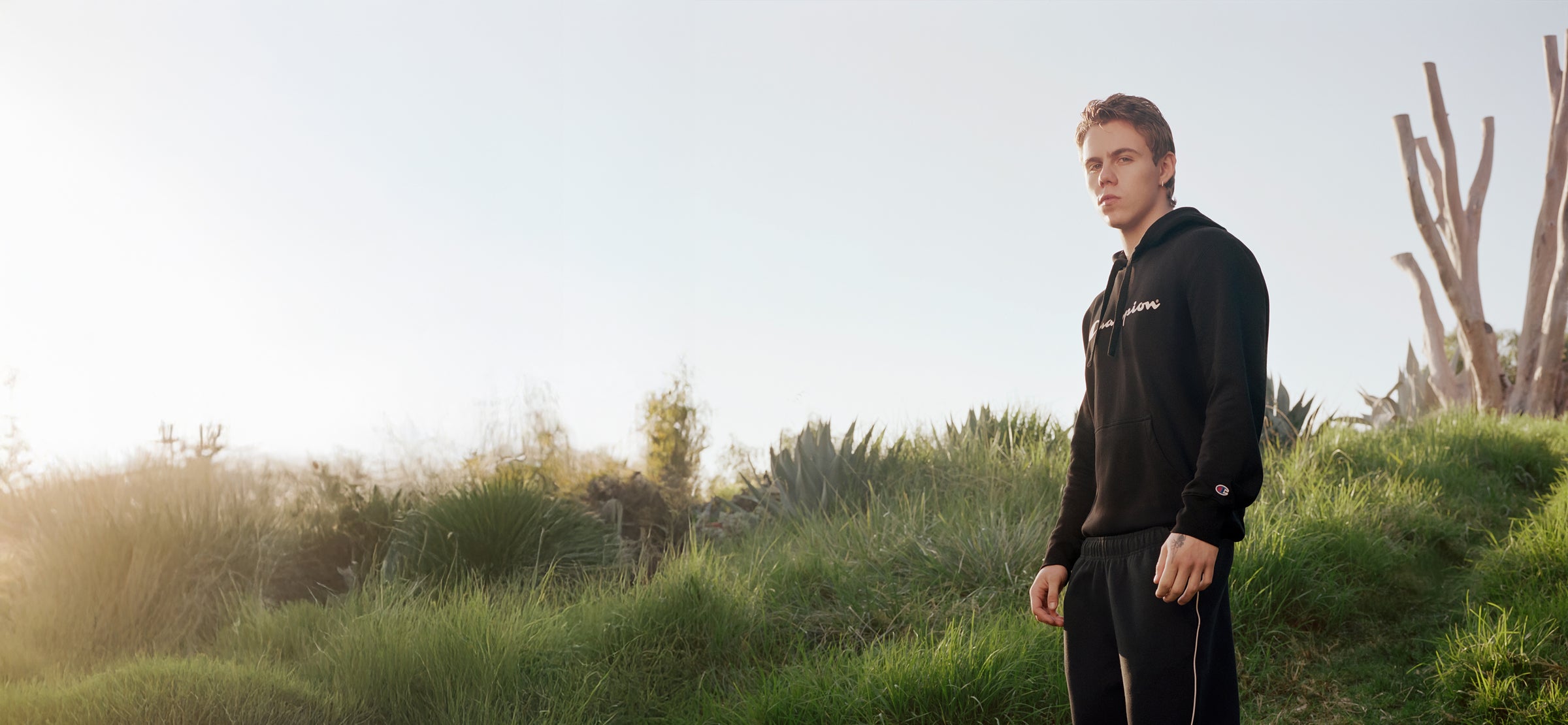 A young man in a black hoodie and sweatpants stands outdoors on green grass with bushes and trees in the background, looking into the distance under a clear, bright sky.