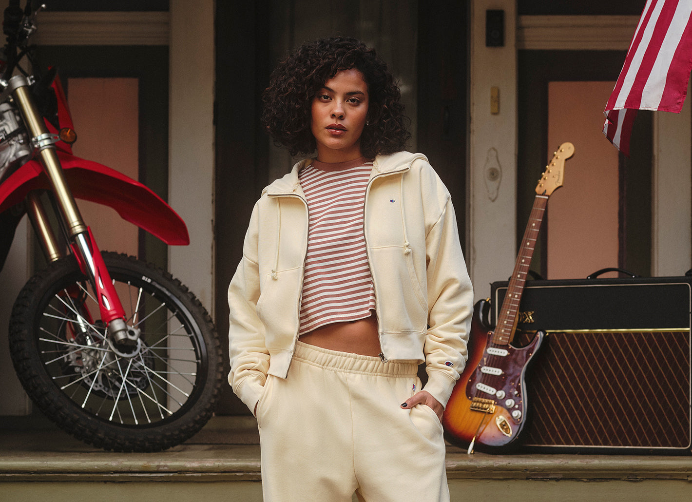 Woman standing in front of a red motorcycle and guitar with an amplifier on a porch.
