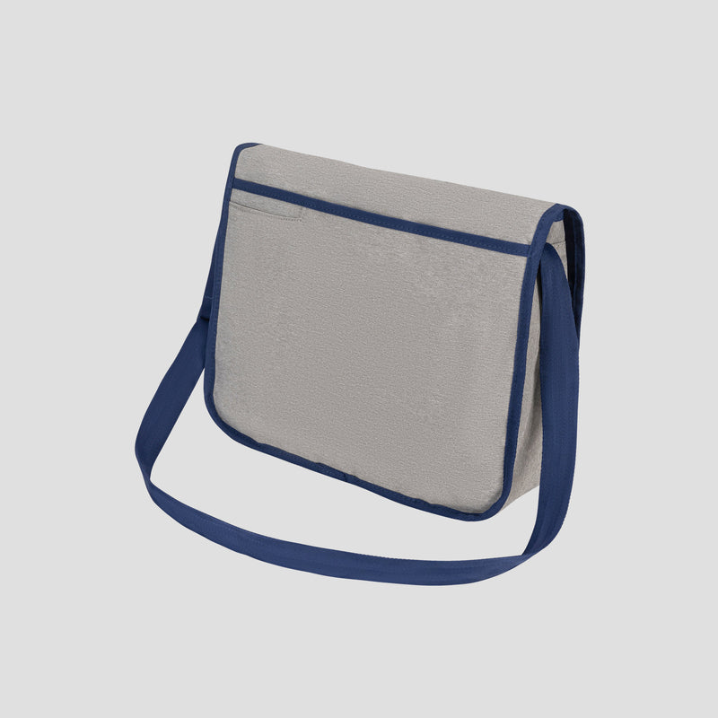 The Champion Phase Messenger Bag, featuring a light gray body with dark blue trim and a matching blue shoulder strap, is showcased against a plain light background. Designed with urban commuters in mind, this bag offers both style and functionality with its flap closure and padded laptop sleeve for added protection on the go.