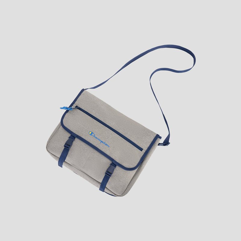 Introducing the Champion Phase Messenger Bag: a gray messenger bag accented with blue trim and a blue shoulder strap, featuring two front buckles, a front zipper pocket, and a small embroidered Champion logo on the front flap. Designed for urban commuters, it also includes a padded laptop sleeve for added convenience.