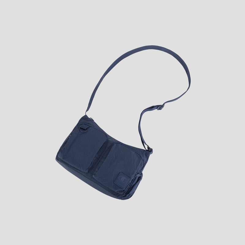 The Quartz Small Messenger Bag by Champion, crafted from lightweight navy blue nylon and featuring two front pockets and an adjustable strap, is displayed against a plain light gray background.