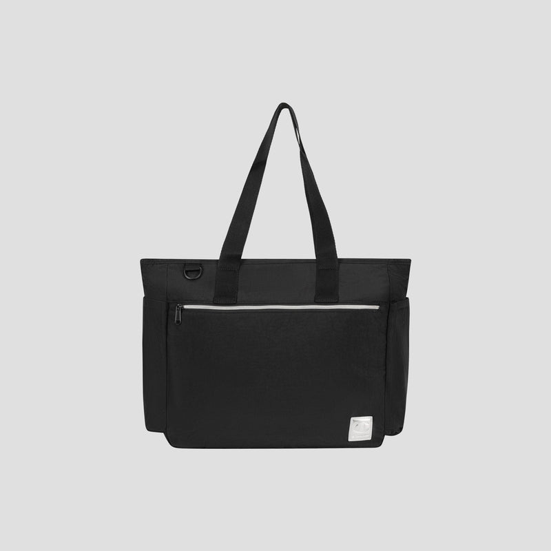 The Champion Quartz East West Tote is a black crinkle nylon bag featuring two shoulder straps, a spacious front zip pocket, side pockets, and a small white label at the bottom right corner. It also includes a padded laptop sleeve and is displayed against a plain light gray background.