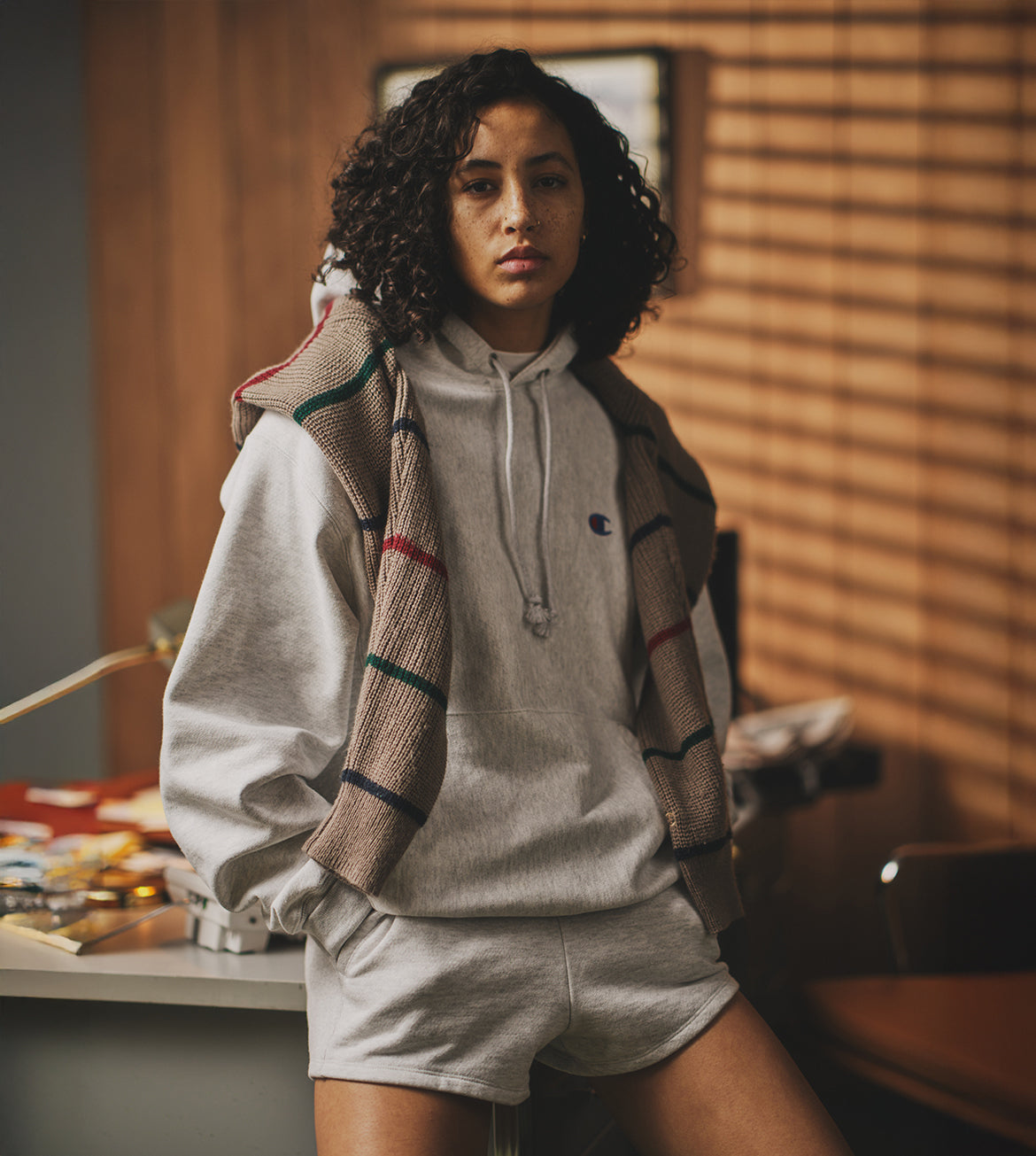 Woman wearing a gray hoodie and beige shorts with a plaid scarf in a room with wooden walls.