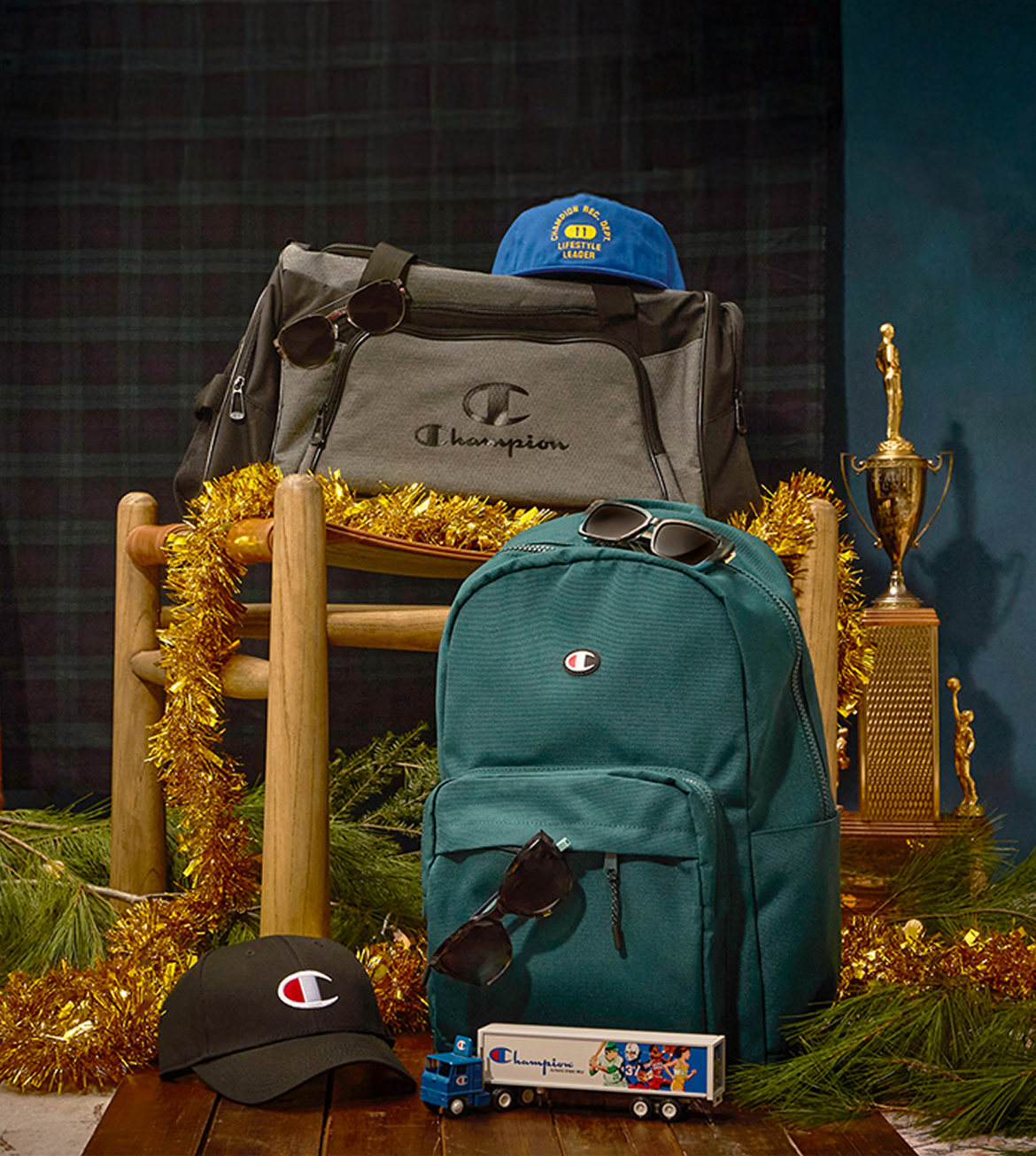 Two Champion backpacks, a blue cap, sunglasses, and a toy truck on a decorative setup with tinsel and a trophy.