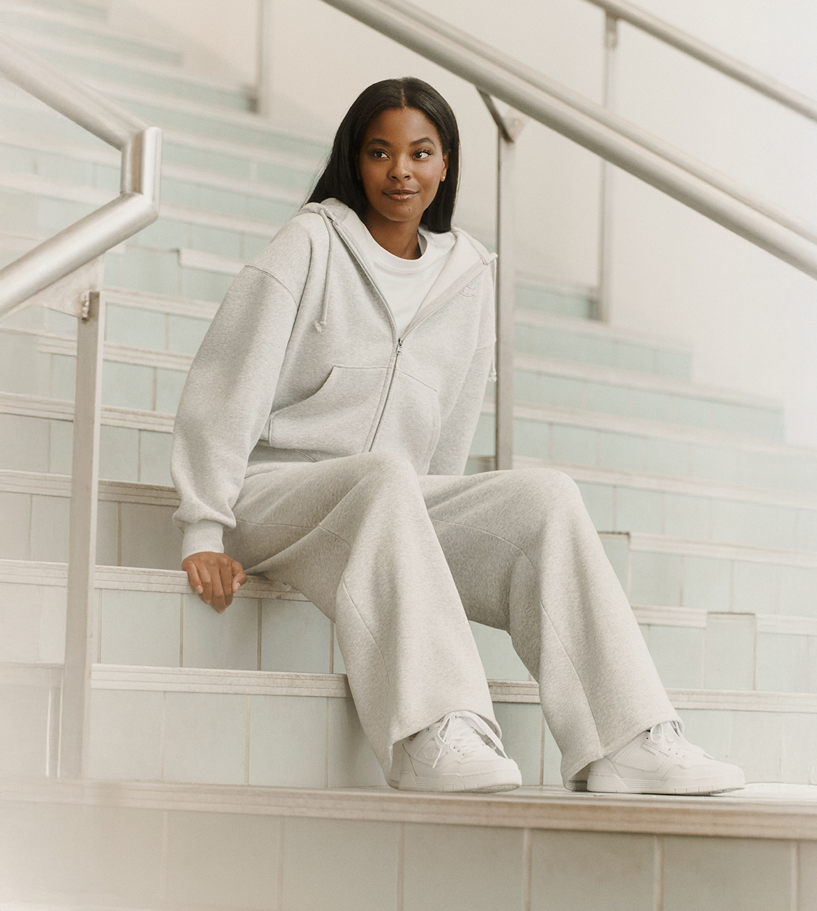 A woman sits on light-colored tiled stairs wearing a matching light gray zip-up hoodie and wide-leg sweatpants with white sneakers.
