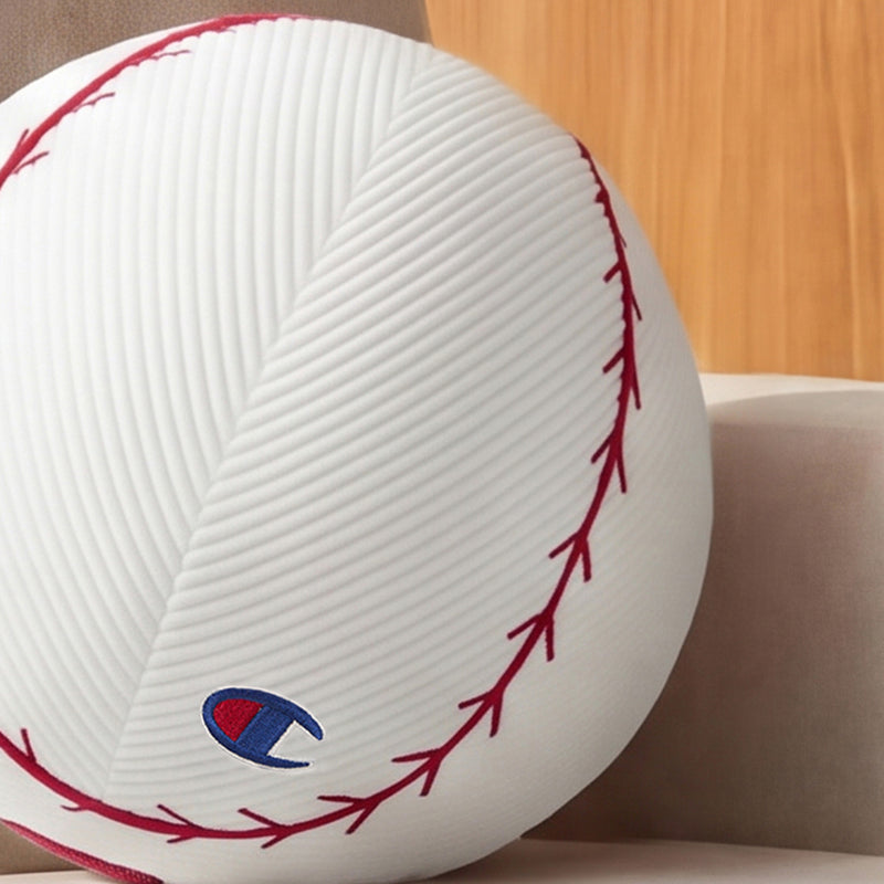 A Champion Sport Shaped Pillow, designed to resemble a baseball with red stitching details and a small blue and red logo, is displayed on a beige surface against a wooden background.