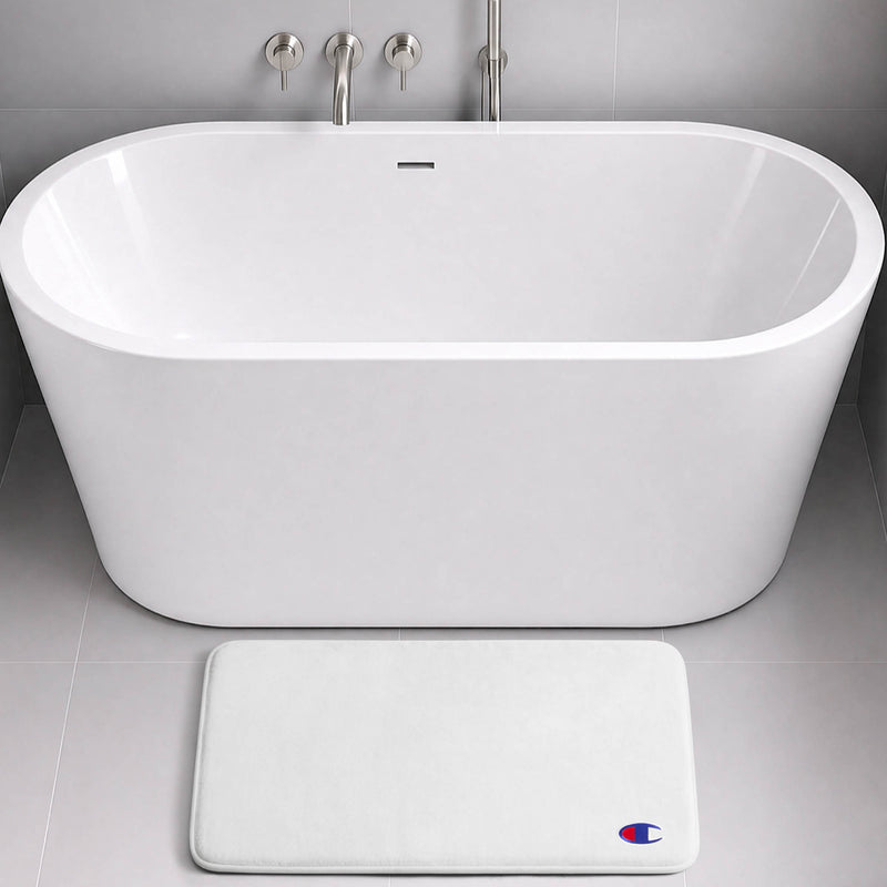 A modern white freestanding bathtub with a wall-mounted faucet sits on gray tile. In front of the tub is the Champion Logo Patch Memory Foam Bath Mat—a white, rectangular bath mat crafted from memory foam for extra comfort and designed to be skid-resistant. A small blue and red Champion logo patch adds a subtle yet stylish touch.