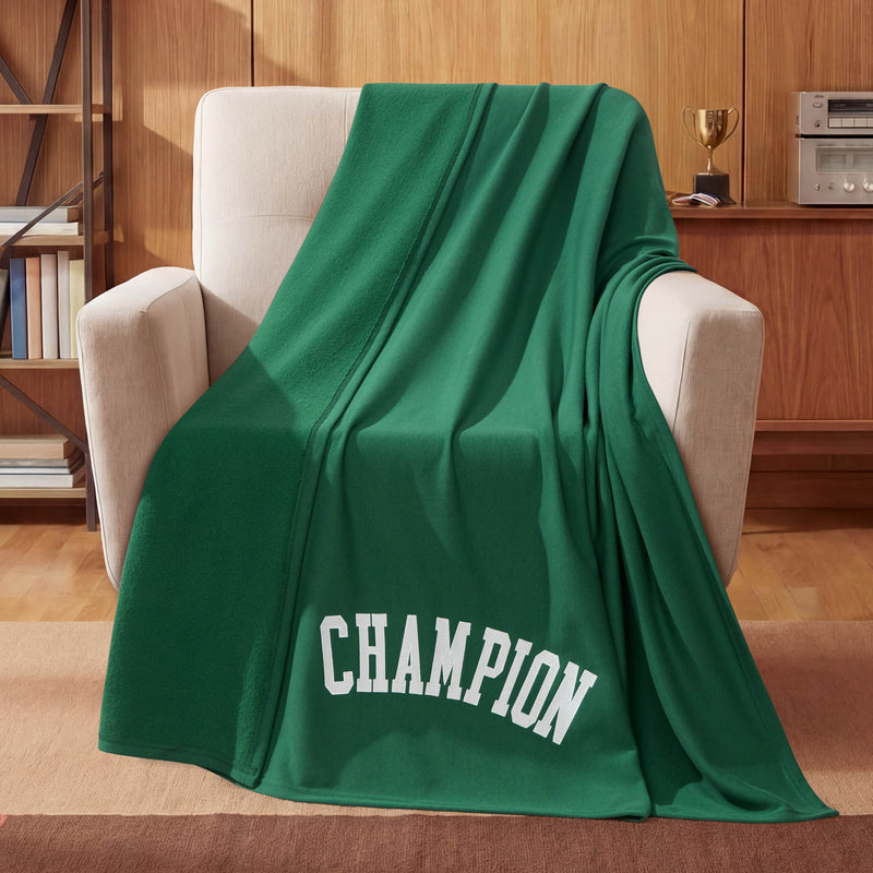 A green Champion Jersey with Fleece Backing Throw Blanket featuring the word CHAMPION in white letters is draped over a beige armchair. This cozy throw from Champion adds a sporty touch to the space, while a small trophy sits on a wooden cabinet in the background.