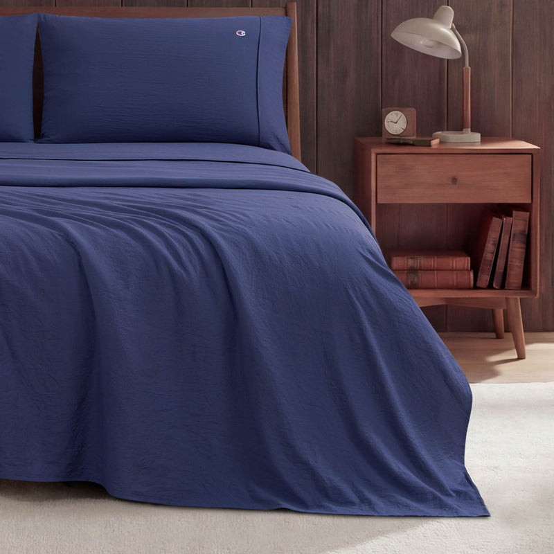A neatly made bed with deep pocket sheets from the Champion Peak Comfort Microfiber Sheet Set in blue, complemented by matching pillowcases, sits next to a wooden nightstand holding a lamp, clock, and books, all set against a wooden wall.