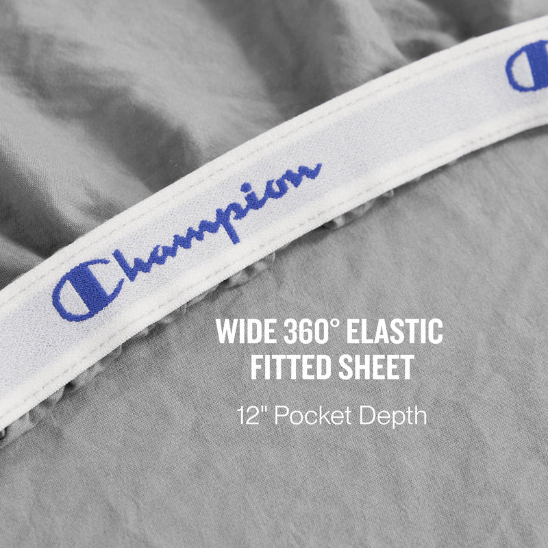 Close-up of a gray fitted sheet featuring a white elastic band labeled Champion. Text reads: Wide 360° elastic fitted sheet, 12″ deep pocket sheets. Part of the Peak Comfort Microfiber Sheet Set by Champion—athletic bedding designed for superior fit and comfort.
