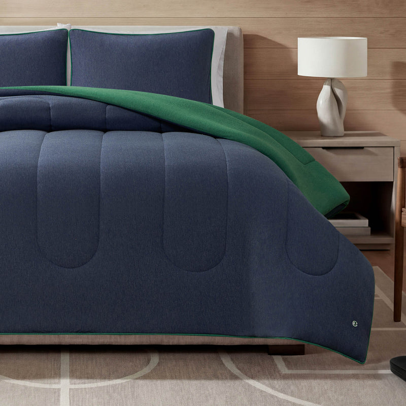 A bed featuring the Champion Premium Reversible Solid Jersey Comforter Set in navy blue and green, paired with matching pillows, alongside a wooden nightstand topped with a white lamp, and a light-colored rug adorned with a geometric pattern.