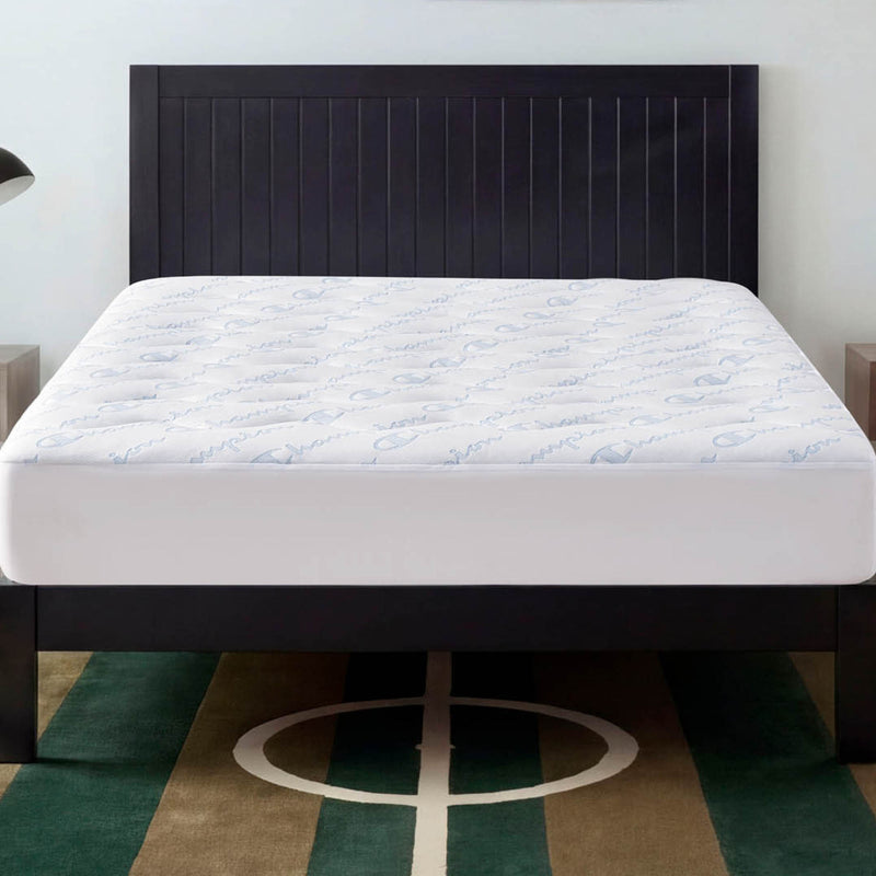 A neatly made bed with a white, antimicrobial and stain-resistant mattress is topped by the Champion Cooling Knit Mattress Pad and features a dark wooden headboard, all situated on a green and brown rug with a white geometric pattern.