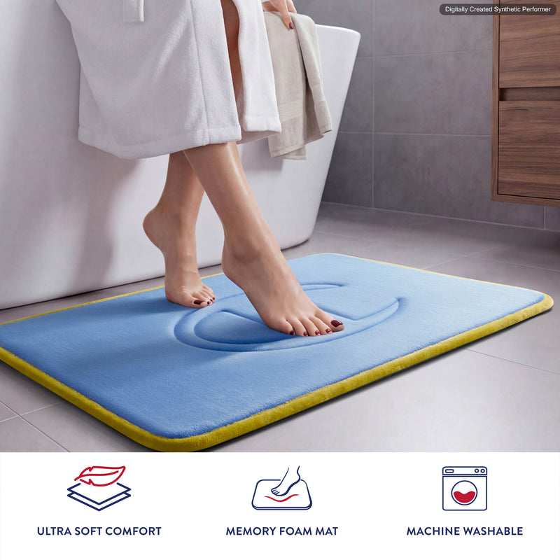 A person in a white robe steps onto a plush blue Champion Quilted Logo Memory Foam Bath Mat near a bathtub. Icons below highlight features: ultra-soft comfort, memory foam support, and machine washable design.