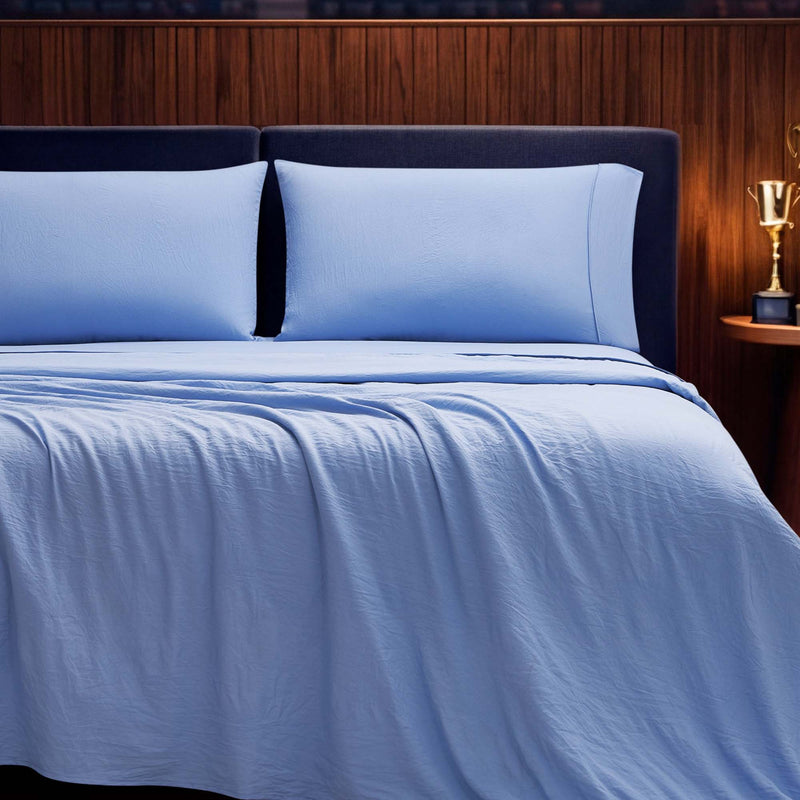 A neatly made bed showcases the Champion Elastic Logo Microfiber Sheet Set in light blue, featuring the distinctive Champion elastic logo detail along the edges and two matching pillows, all set against a dark wood-paneled headboard with a nightstand displaying a golden trophy.