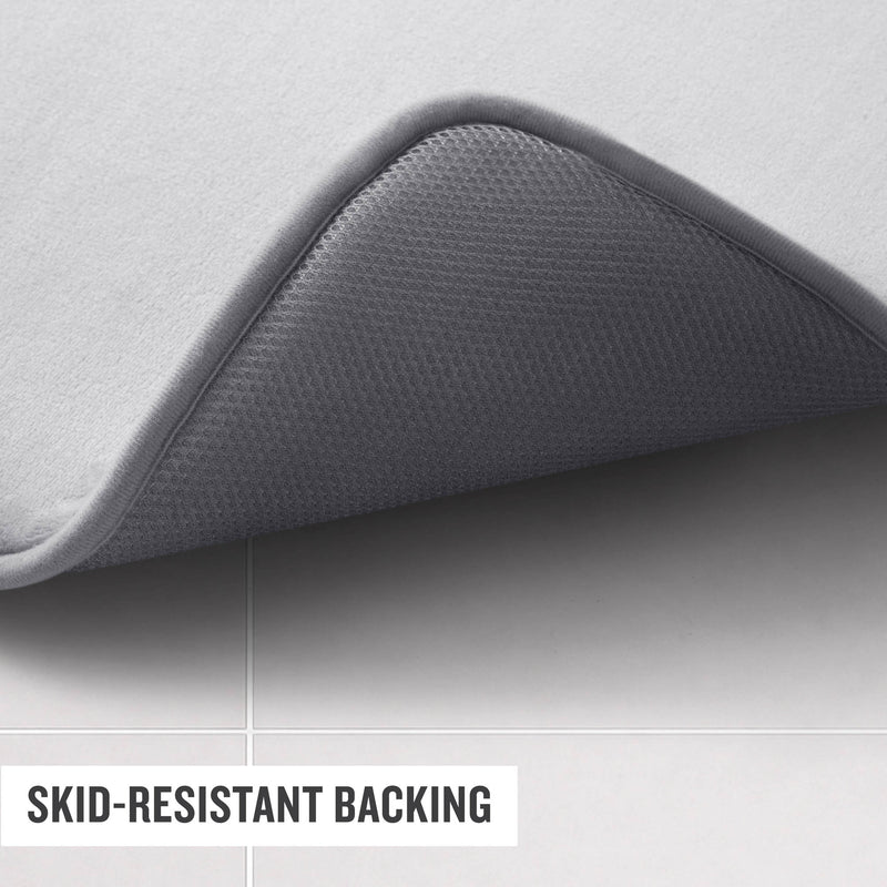 A Champion Logo Patch Memory Foam Bath Mat in gray is displayed with one corner lifted to reveal its textured, skid-resistant backing. The mat rests on light-colored tiles, and text on the image reads “SKID-RESISTANT BACKING.”.