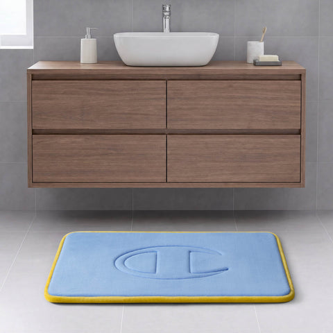 A modern bathroom with a floating wooden cabinet, white sink, soap dish, and a Champion Quilted Logo Memory Foam Bath Mat in blue with a yellow border and embossed design on a light gray tiled floor.