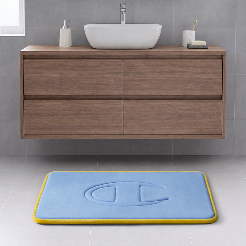 A modern bathroom with a floating wooden cabinet, white sink, soap dish, and a Champion Quilted Logo Memory Foam Bath Mat in blue with a yellow border and embossed design on a light gray tiled floor.