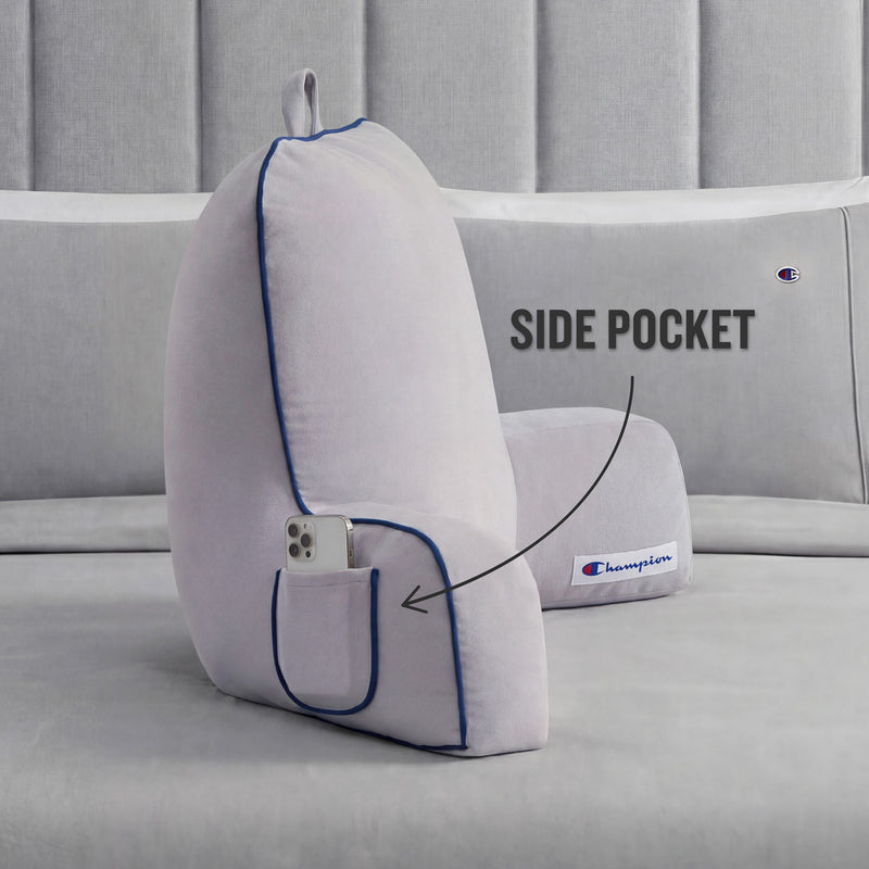 A light gray Champion Faux Wool Backrest Pillow with supportive arms and blue piping rests on a gray bed. Made from soft faux wool, the pillow includes a small side pocket—highlighted with an arrow and the label “SIDE POCKET”—holding a smartphone. The Champion logo patch is also visible.