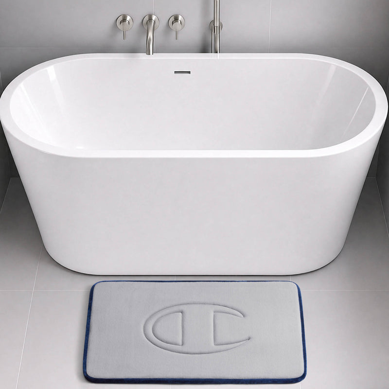 A modern white bathtub with wall-mounted faucets sits in a grey tiled bathroom. In front of the tub is a light grey Champion Quilted Logo Memory Foam Bath Mat, featuring a raised C logo, plush memory foam comfort, and a dark blue border.
