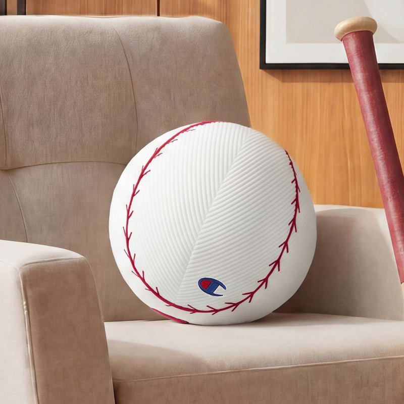 A large, round Champion Sport Shaped Pillow designed to resemble a baseball rests on a beige armchair, with part of a wooden baseball bat visible nearby.