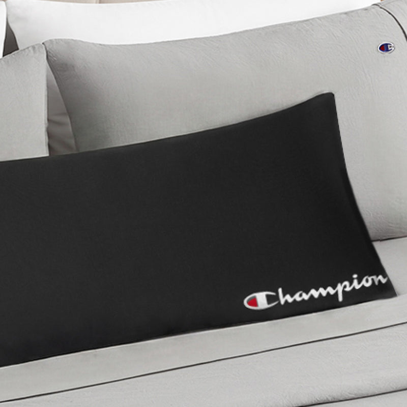 A black Champion Jersey Body Pillow featuring the iconic logo and script rests on a bed dressed in light gray pillows and sheets adorned with small Champion logos, creating a sporty, athletic accent.