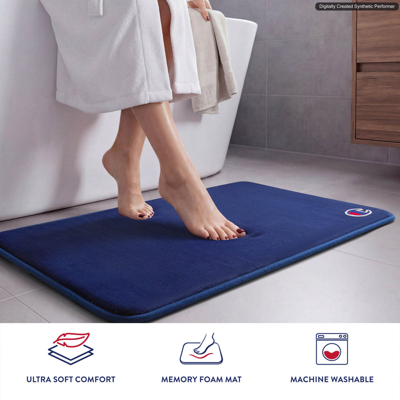 A person in a white robe steps onto the Champion Logo Patch Memory Foam Bath Mat, enjoying the ultra-soft comfort of its skid-resistant, blue memory foam design in a modern bathroom. Three icons below highlight its features: ultra soft comfort, memory foam mat, and machine washable.