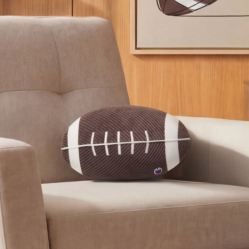 A Champion Sport Shaped Pillow, crafted to resemble a football with white laces, rests on a beige armchair in a wood-paneled room adorned with a framed picture above.