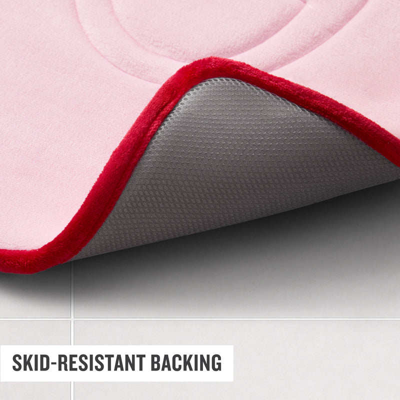 A close-up of the Champion Quilted Logo Memory Foam Bath Mat in pink with a red border, highlighting its gray skid-resistant backing, placed on a light tiled floor. Text reads SKID-RESISTANT BACKING.