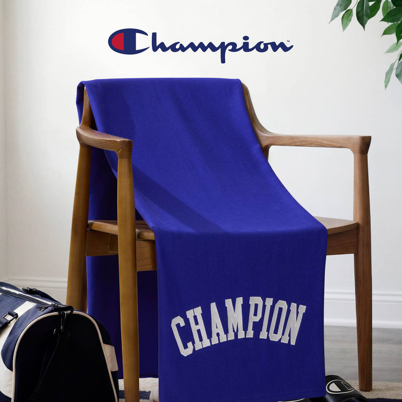 A Champion Jersey with Fleece Backing Throw Blanket in blue, featuring the CHAMPION name in white letters, is draped over a wooden chair. The Champion logo appears on the wall behind, and a navy-and-white gym bag is partially visible on the floor.
