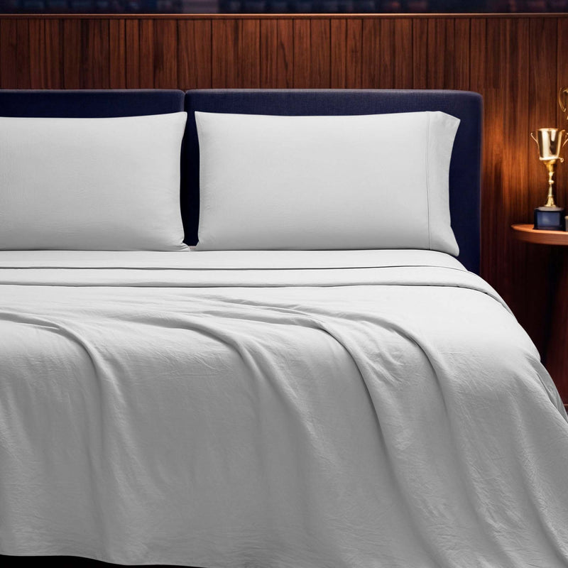 A neatly made bed with crisp white Champion Elastic Logo Microfiber Sheet Set and pillows, a blue upholstered headboard, and a wooden nightstand holding a gold trophy, set against a wood-paneled wall.