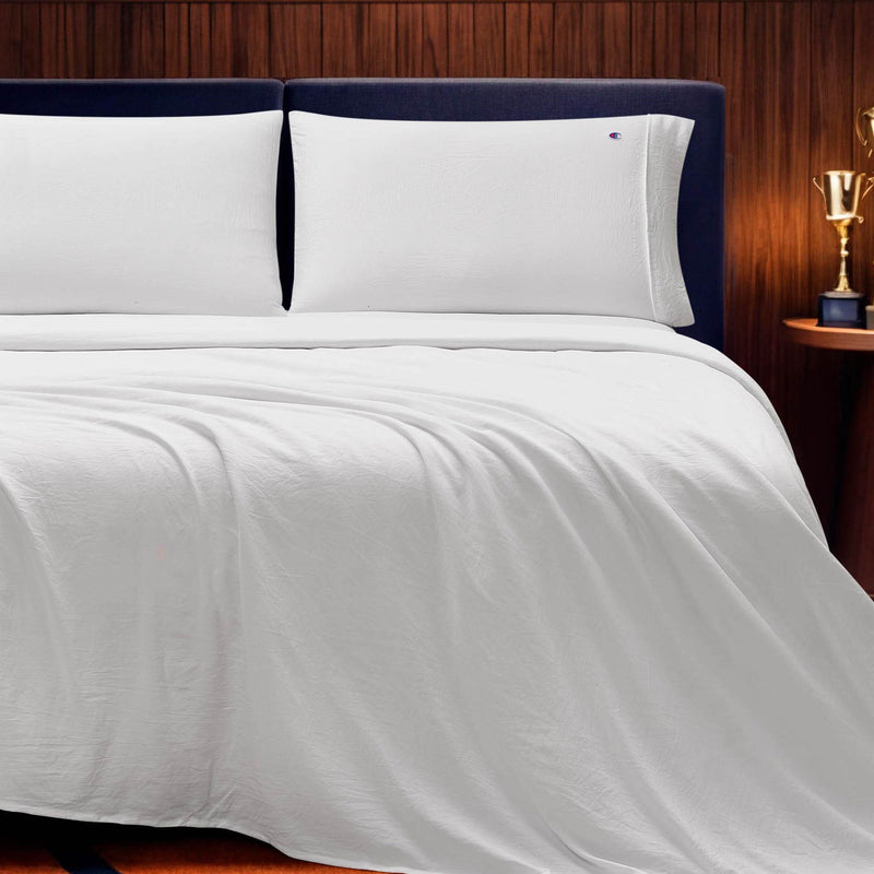 A neatly made bed with crisp white Champion Embroidery Patch Microfiber Sheet Set and pillows is set against a dark headboard and wood-paneled wall. A small table with a gold trophy stands beside the bed, enhancing the athletic-inspired bedding vibe.